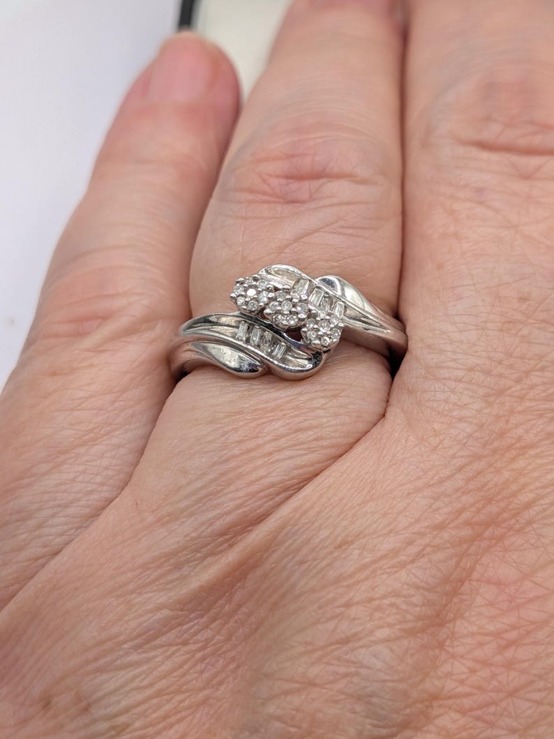May include: A silver ring with a cluster of small round diamonds and baguette-cut diamonds. The ring has a swirling design with the diamonds set in a raised setting. The ring is on a finger.