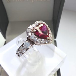 May include: A heart-shaped ruby ring with a halo of smaller diamonds. The ring is set in rose gold and has a polished finish.