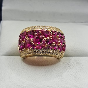 May include: A gold ring with a band of bright pink gemstones, complemented by smaller clear stones. The ring's design features a cluster of pink gems, creating a striking piece of jewellery.