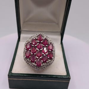 Puede incluir: Un anillo de plata con un grupo de doce rubíes rojos de talla ovalada dispuestos en cuadrícula, rodeados por un borde de pequeñas piedras transparentes, presentado dentro de un joyero verde oscuro con bisagras y forro interior blanco.