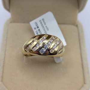 May include: A gold ring with a rounded design, adorned with diagonal rows of clear gemstones. The ring is displayed in a beige jewellery box, with a tag stating "Made with SWAROVSKI".