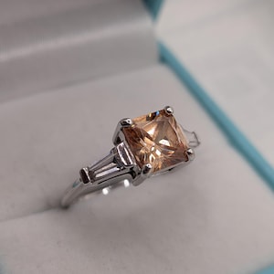 May include: A silver-toned ring featuring a large, square-cut, peach-coloured gemstone at the centre, flanked by two smaller, elongated baguette-cut clear stones on each side, all set within a polished metal band with four prongs per stone.