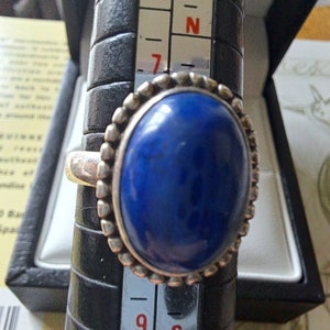 May include: A silver ring with a large, oval-shaped blue gemstone set in a beaded bezel. The ring is on a sizing tool, showing a size 9.