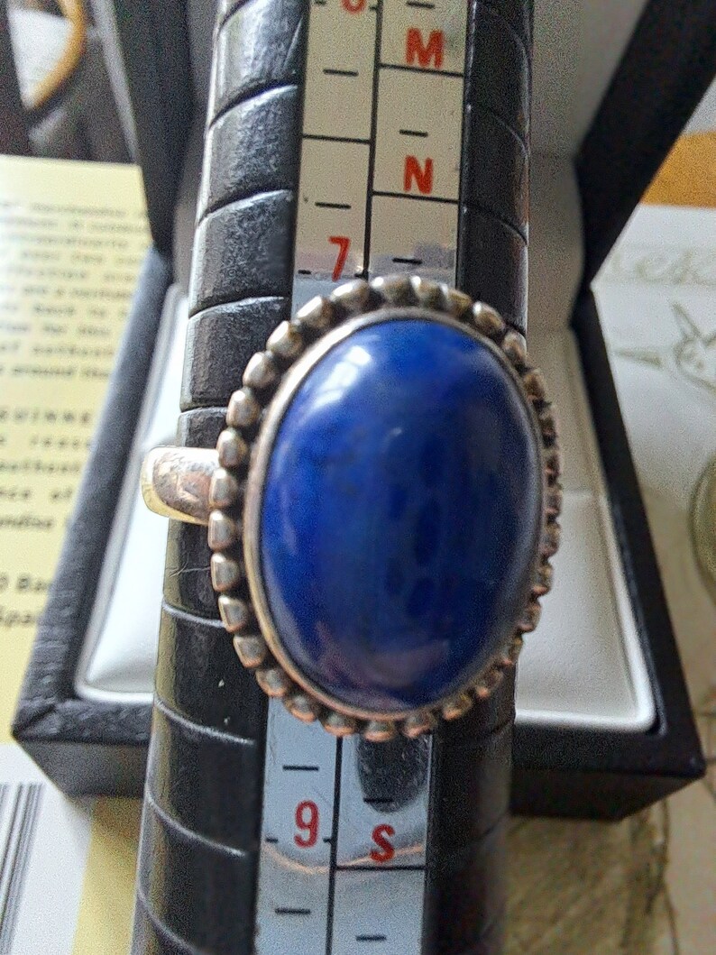 May include: A silver ring with a large, oval-shaped blue gemstone set in a beaded bezel. The ring is on a sizing tool, showing a size 9.