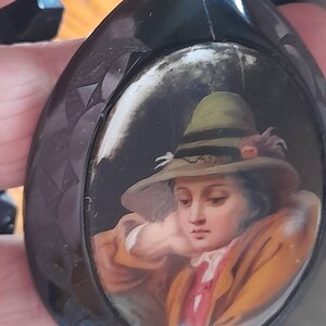 May include: A black oval locket with a painted portrait of a young person wearing a green hat. The portrait is set in a white oval frame.