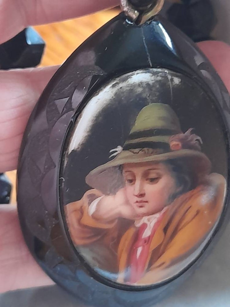May include: A black oval locket with a painted portrait of a young person wearing a green hat. The portrait is set in a white oval frame.