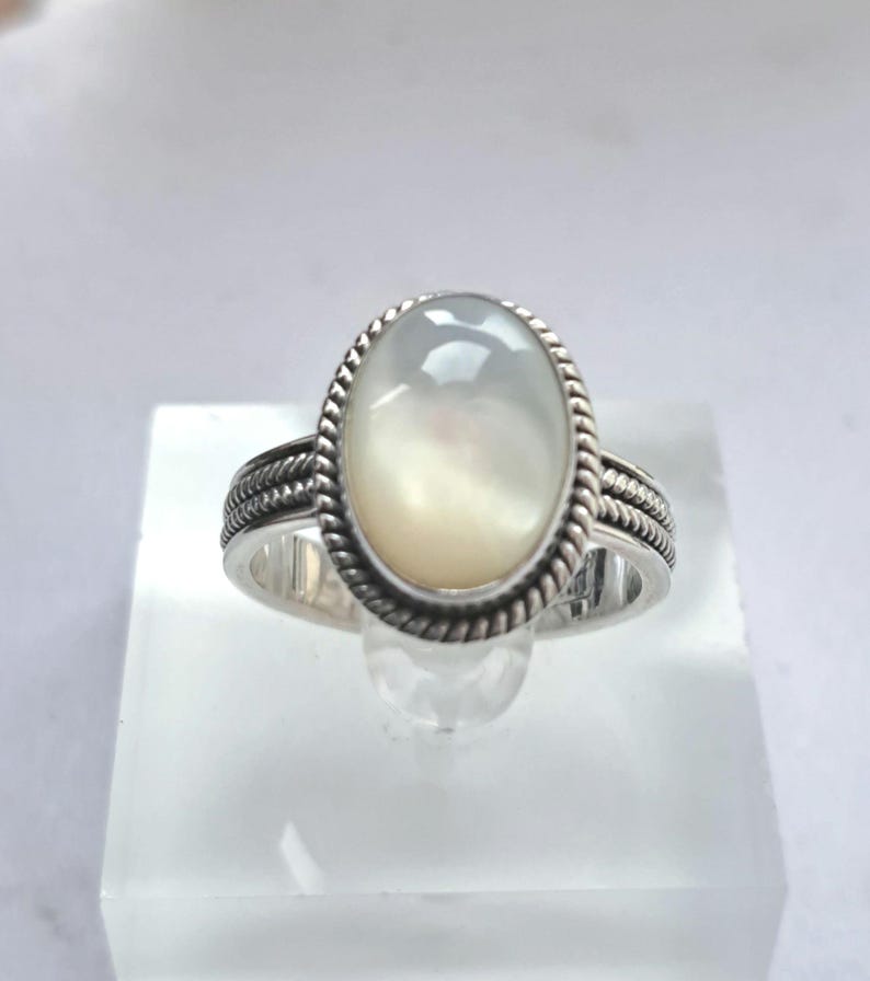 May include: A silver ring with a white oval mother of pearl gemstone. The ring has a rope-like design around the band and the gemstone.