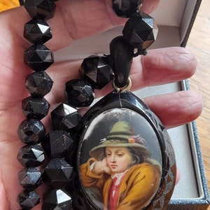 May include: A black jet necklace with a black oval pendant featuring a painted portrait of a young person wearing a green hat. The pendant has a detailed carved design around the edge.