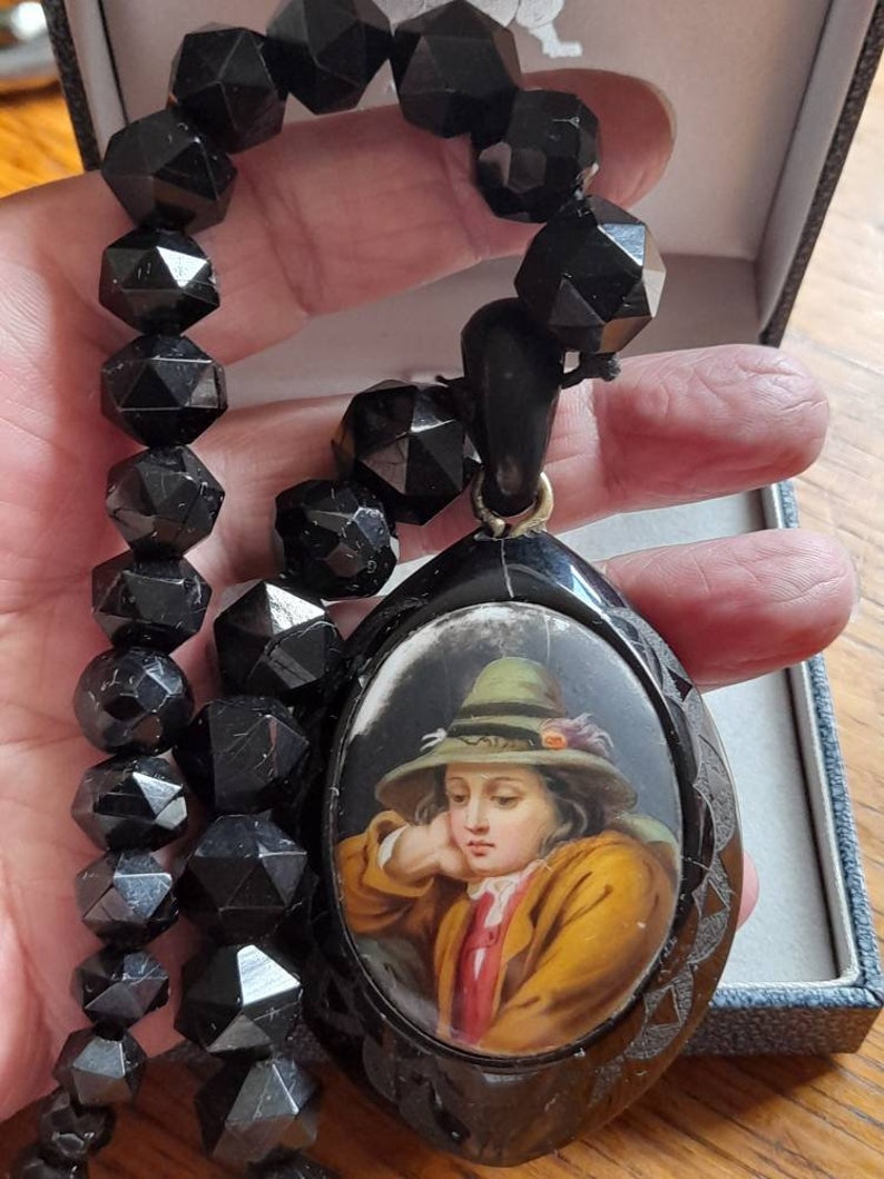 May include: A black jet necklace with a black oval pendant featuring a painted portrait of a young person wearing a green hat. The pendant has a detailed carved design around the edge.