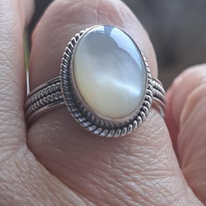 May include: A silver ring with a white oval gemstone set in a rope-like design.
