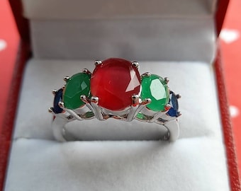 May include: A silver ring with three oval-shaped gemstones. The center stone is red, and the two flanking stones are green. The ring is in a white box.