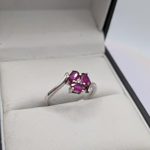 Può includere: Un anello d'argento con un gruppo di pietre preziose rosa scuro a taglio ovale, accentuato da una piccola pietra preziosa trasparente al centro. L'anello è incastonato in una fascia d'argento ed è esposto in un cofanetto nero e bianco.