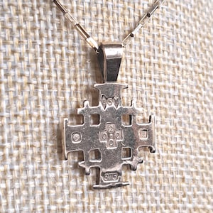 Puede incluir: Collar con colgante plateado con diseño de cruz de Jerusalén. El colgante tiene un patrón calado detallado con pequeños recortes cuadrados y está suspendido de una delicada cadena. El colgante está marcado con el número "375".