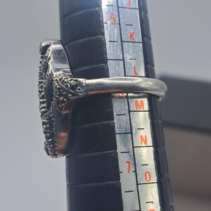 May include: A silver ring with a textured oval design, set on a black ring sizer. The ring's band is smooth, and the oval is filled with small, dark stones. The ring is being measured for size.