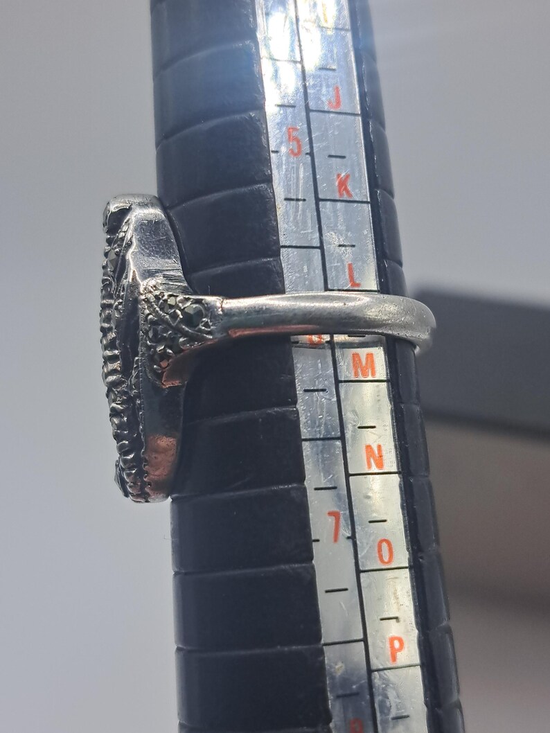 May include: A silver ring with a textured oval design, set on a black ring sizer. The ring's band is smooth, and the oval is filled with small, dark stones. The ring is being measured for size.