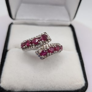May include: A bypass style ring featuring two parallel rows of oval-cut deep red rubies set in a silver-toned metal band. Each row is accented with small, clear pavé stones along the edges, creating a sparkling, layered appearance on the finger.
