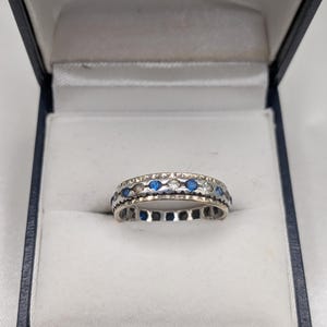 May include: Silver ring with alternating blue and clear stones. The ring is set in a dark blue jewelry box with a white satin interior. The box has a gold logo on the inside of the lid.