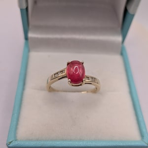 Puede incluir: Un anillo de oro con una piedra preciosa rosa de corte cabujón ovalado en el centro, sujeta por cuatro garras. La banda está decorada con pequeñas piedras claras engastadas en canal a ambos lados, presentado en un joyero azul claro.