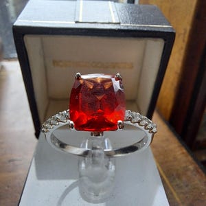 Peut inclure: Une bague en argent ornée d'une grosse pierre rouge vif de taille carrée. La monture est ornée de petites pierres transparentes. La bague est présentée sur un présentoir transparent, avec une boîte noire en arrière-plan. La boîte porte l'inscription "NORTHERN COUNTRIES" en lettres dorées.