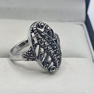 May include: A silver-toned ring with an oval filigree design. The ring features small, dark stones set within the filigree pattern. The ring is displayed on a white surface.