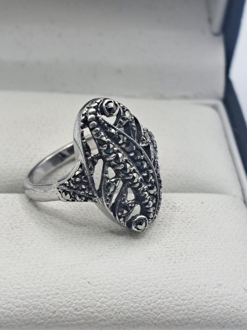May include: A silver-toned ring with an oval filigree design. The ring features small, dark stones set within the filigree pattern. The ring is displayed on a white surface.