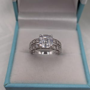 May include: A silver-toned ring featuring a large, rectangular, emerald-cut clear centre stone held by four prongs. The band is split into two rows, each channel-set with multiple small, round, clear stones, creating a wide, multi-faceted appearance.