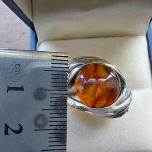 May include: A silver ring with a large amber stone set in the center. The ring is in a blue velvet box with a ruler for size reference.