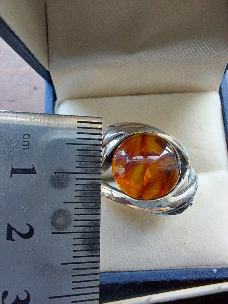 May include: A silver ring with a large amber stone set in the center. The ring is in a blue velvet box with a ruler for size reference.