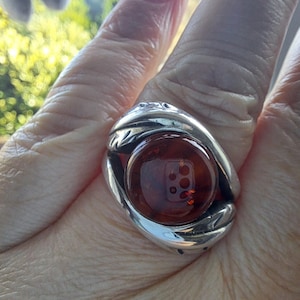 May include: A silver ring with a large, round, amber stone set in the center. The ring has a detailed design around the stone.