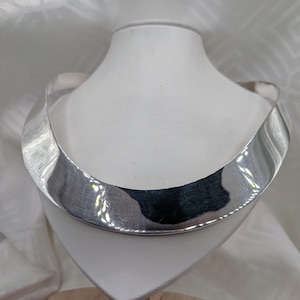 Könnte beinhalten: Eine breite, polierte silberfarbene Halsspange mit einer glatten, geschwungenen und reflektierenden Oberfläche. Die Halskette zeichnet sich durch ein starres, offenes Design aus, das flach am Hals anliegt und ein glänzendes Finish aufweist.