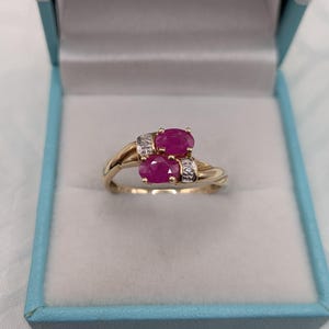 May include: A gold ring featuring two oval-cut vibrant pink rubies set in a bypass style. Each ruby is accented by a small, clear, round-cut diamond set into the gold band on either side of the stones, creating a balanced and elegant design.