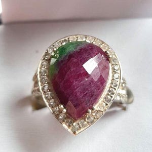 May include: A silver ring with a pear-shaped, faceted ruby gemstone and a green section. The ruby is encircled by small, clear gemstones. The ring's band has a textured design.