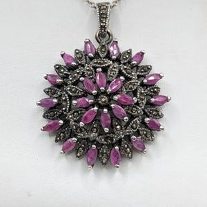 May include: A silver pendant necklace with a circular pendant. The pendant showcases oval, deep pink gemstones and smaller, dark grey stones, arranged in a floral design. The necklace is completed with a fine silver chain.