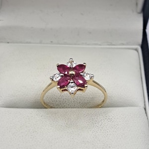 Könnte beinhalten: Ein goldener Ring mit einem Blumenmuster. Die Blume besteht aus roten und weißen Edelsteinen. Der Ring hat ein goldenes Band und wird in einer weißen Schmuckschatulle präsentiert.