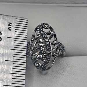 May include: A silver-toned filigree ring with an oval design. The ring features intricate openwork and is embellished with small, dark stones. The ring's length is approximately 1.25 inches.