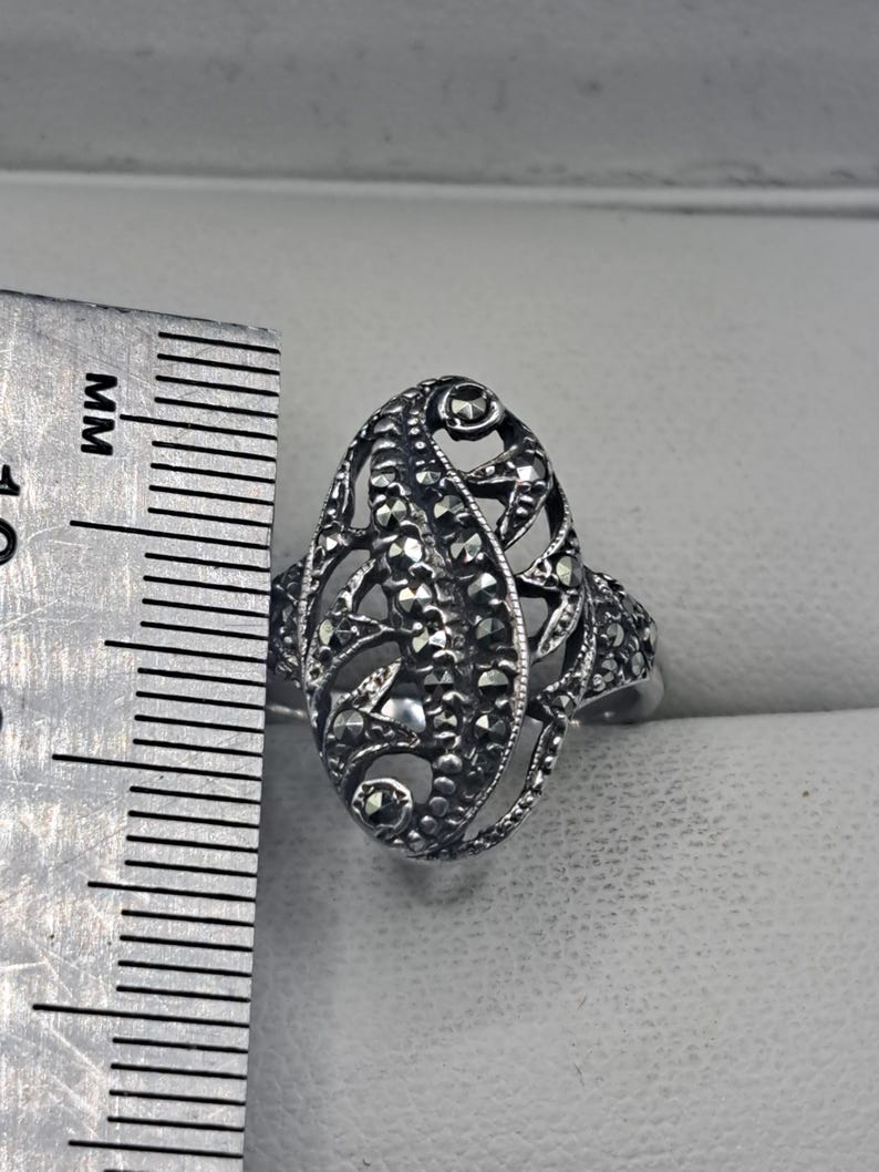 May include: A silver-toned filigree ring with an oval design. The ring features intricate openwork and is embellished with small, dark stones. The ring's length is approximately 1.25 inches.