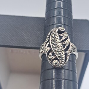 May include: A silver ring with an oval marcasite design. The ring features a central marcasite-studded section with a leaf-like pattern, surrounded by smaller marcasite accents. The ring is displayed on a black ring sizer.