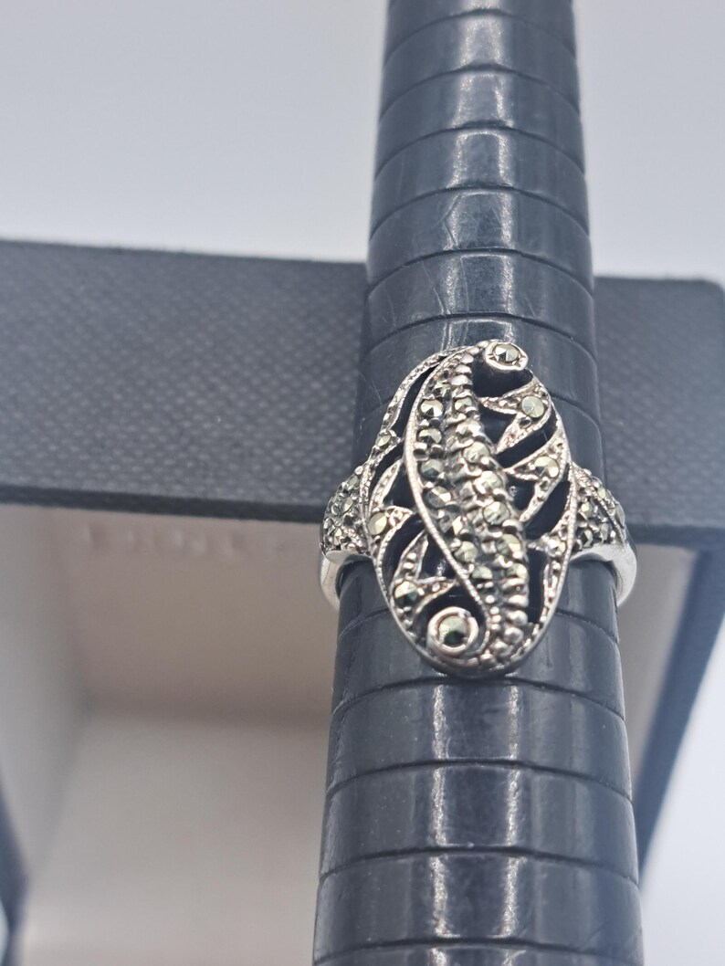May include: A silver ring with an oval marcasite design. The ring features a central marcasite-studded section with a leaf-like pattern, surrounded by smaller marcasite accents. The ring is displayed on a black ring sizer.
