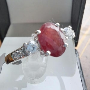 May include: A silver ring featuring a large oval red gemstone, surrounded by smaller clear gemstones.