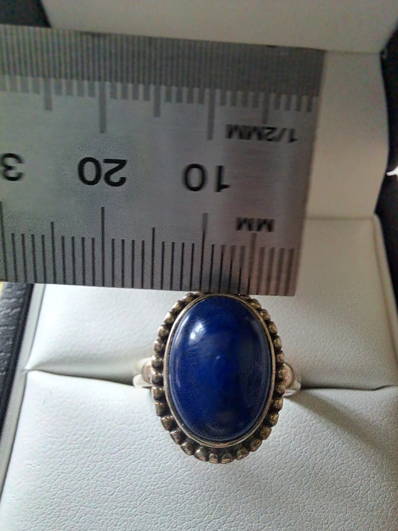 May include: A silver ring with a dark blue oval gemstone set in a beaded bezel. The ring is measured against a ruler.