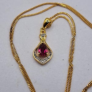 May include: A gold necklace with a pendant. The pendant has a teardrop-shaped, deep red gemstone, set in a gold frame with small, clear accent stones. The chain is a delicate, textured design.