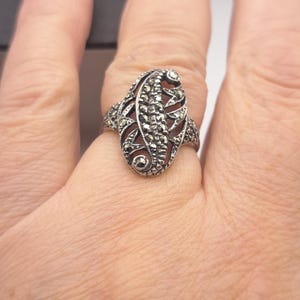 May include: A silver-toned ring with an oval, ornate design. The ring features a central leaf-like pattern with small, sparkling accents. The ring is worn on a finger, showcasing its intricate details and vintage style.