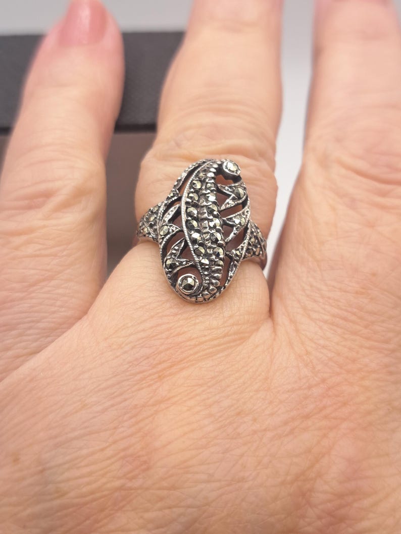 May include: A silver-toned ring with an oval, ornate design. The ring features a central leaf-like pattern with small, sparkling accents. The ring is worn on a finger, showcasing its intricate details and vintage style.