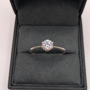 Peut inclure: Une bague de fiançailles solitaire en argent avec un diamant rond. La bague est présentée dans un écrin en velours noir, mettant en valeur l'éclat du diamant. Un bijou intemporel, symbole classique d'amour et d'engagement.