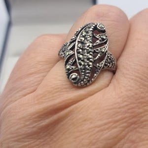 May include: A silver-toned ring with an elongated oval design. The ring features a detailed pattern of small, sparkling stones and openwork filigree, creating a vintage or antique aesthetic. The ring is worn on a finger.