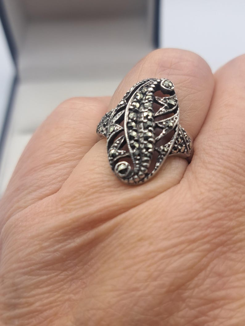 May include: A silver-toned ring with an elongated oval design. The ring features a detailed pattern of small, sparkling stones and openwork filigree, creating a vintage or antique aesthetic. The ring is worn on a finger.