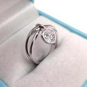 Op de afbeelding: Een zilverkleurige ring met een gespleten bandontwerp en een bungelende hartvormige bedel met een heldere edelsteen. De ring is vervaardigd met een gepolijste metalen afwerking en een enkele gefacetteerde steen in een hartvormig frame.
