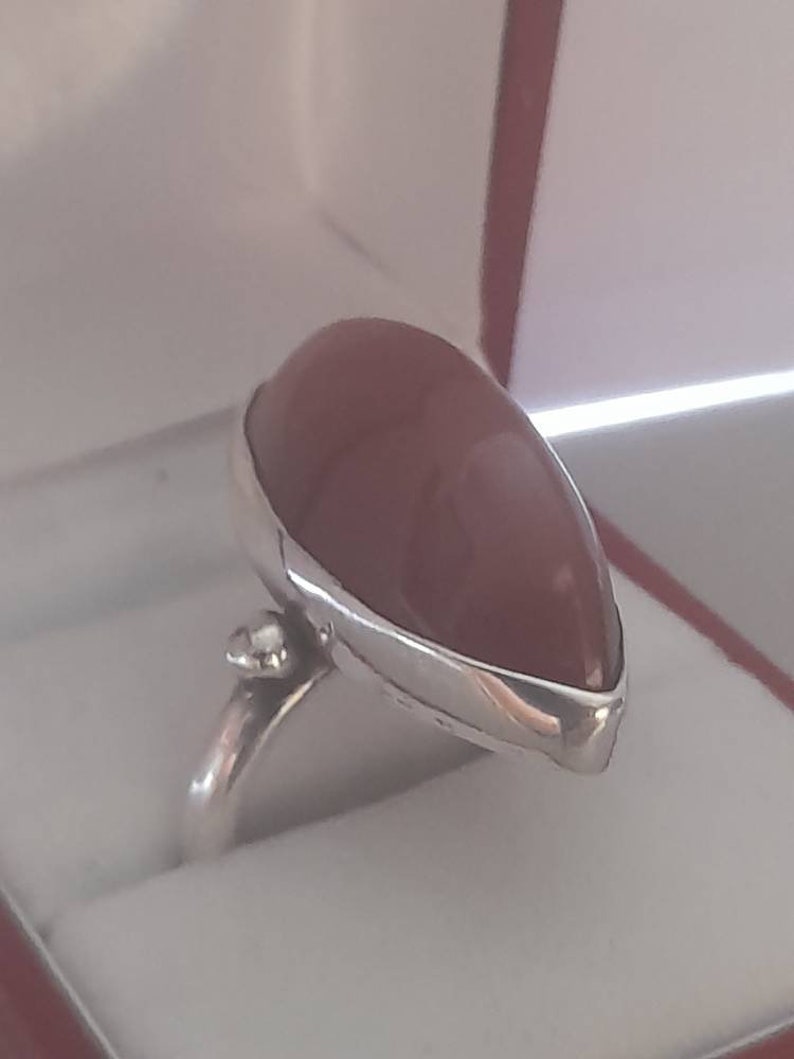 May include: A silver ring with a teardrop-shaped, reddish-brown gemstone. The ring is set in a silver band with a small, clear stone on the side.
