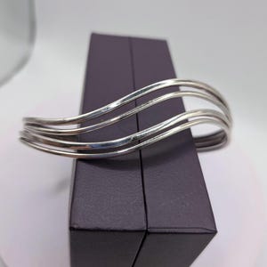Puede incluir: Una pulsera tipo brazalete de color plateado que presenta tres bandas paralelas, onduladas y curvas que crean una apariencia fluida y superpuesta. El metal tiene un acabado pulido y reflectante, manteniendo una forma abierta.