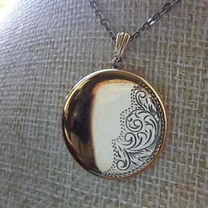 May include: A gold-coloured, round locket necklace with an engraved floral pattern on one side. The locket is suspended from a dark chain. The background is a neutral fabric, accentuating the jewellery's details.